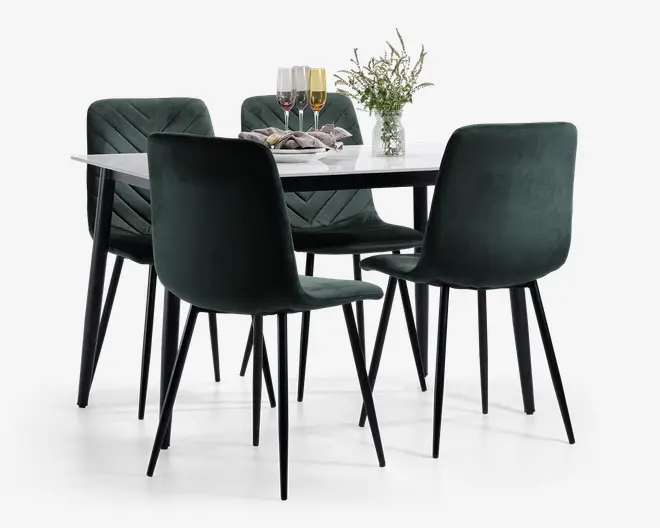 Et moderne spisesæt med et rektangulært hvidt bordplade med sorte ben og fire stole betrukket med mørkegrøn velour. Stolene har bløde, polstrede sæder og ryglæn med diskrete chevron-syninger og slanke, sorte metalben. Bordet er pyntet med en vase med friske blomster, tre vinglas og pænt foldede servietter, hvilket skaber en indbydende og elegant spiseatmosfære.