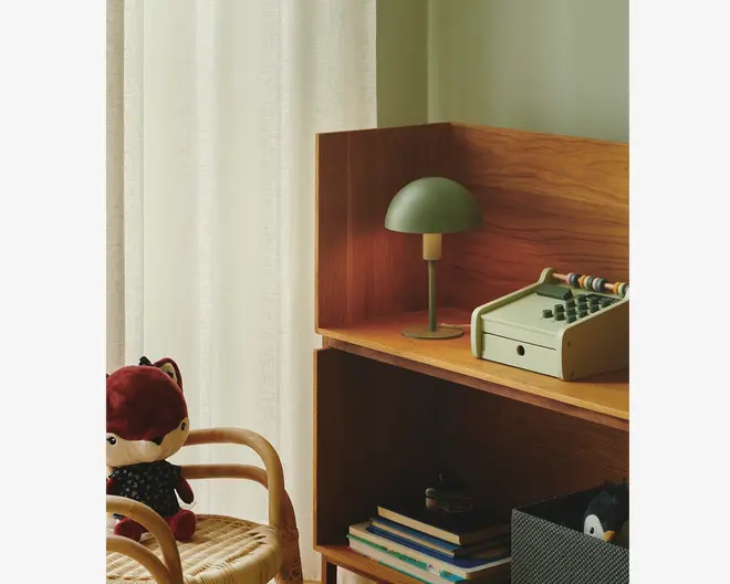 En reol i træ med naturfinish, hvorpå der står en lille grøn bordlampe med kuppelformet skærm, et retro-inspireret legetøjskasseapparat i afdæmpet grøn med farverige kugler samt en stak bøger på nederste hylde. Til venstre ses en rattanstol med en plysbamse formet som en ræv siddende på sædet. Baggrunden består af lyse gardiner og en lysegrøn væg, hvilket skaber en hyggelig og indbydende stemning.