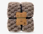 Et sammenfoldet beige tæppe med fluffy, struktureret overflade, bundet med sorte stropper og en brun papiretiket i midten.