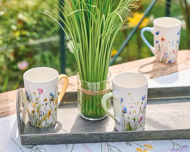 To keramikkrus med blomstereng-motiver, det ene med gult håndtag og det andet med lysegrønt håndtag, står på en rustik metalbakke. Krusene har farverige vilde blomster og græsser trykt på hvid baggrund. Mellem krusene står en klar glasvase fyldt med højt, grønt kunstgræs bundet med sejlgarn. I baggrunden ses endnu et matchende krus med blåt håndtag på et træbord. Omgivelserne er udendørs med en sløret have i baggrunden.