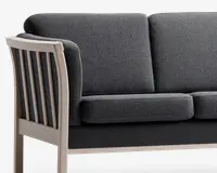 Nærbillede af en sofa med mørkegråt stofbetræk og tykke, rektangulære sæde- og ryghynder. Armlænet og rammen er lavet af lyst træ med lodrette lameller på siden, hvilket giver et moderne og robust udtryk.