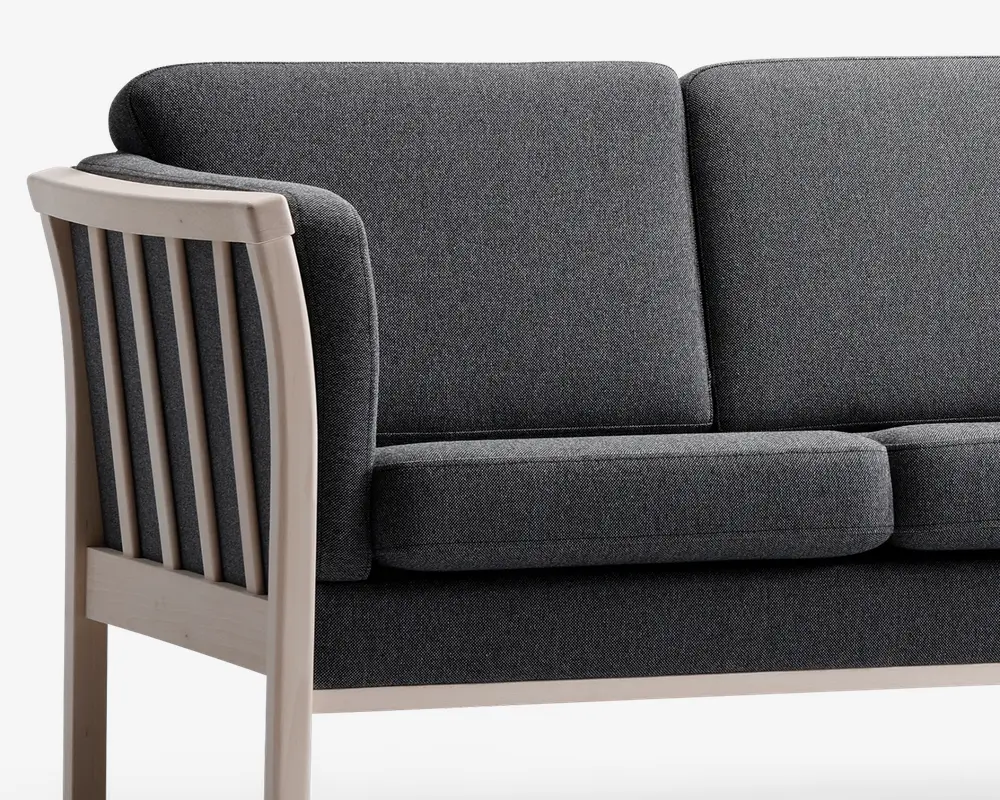 Nærbillede af en sofa med mørkegråt stofbetræk og tykke, rektangulære sæde- og ryghynder. Armlænet og rammen er lavet af lyst træ med lodrette lameller på siden, hvilket giver et moderne og robust udtryk.