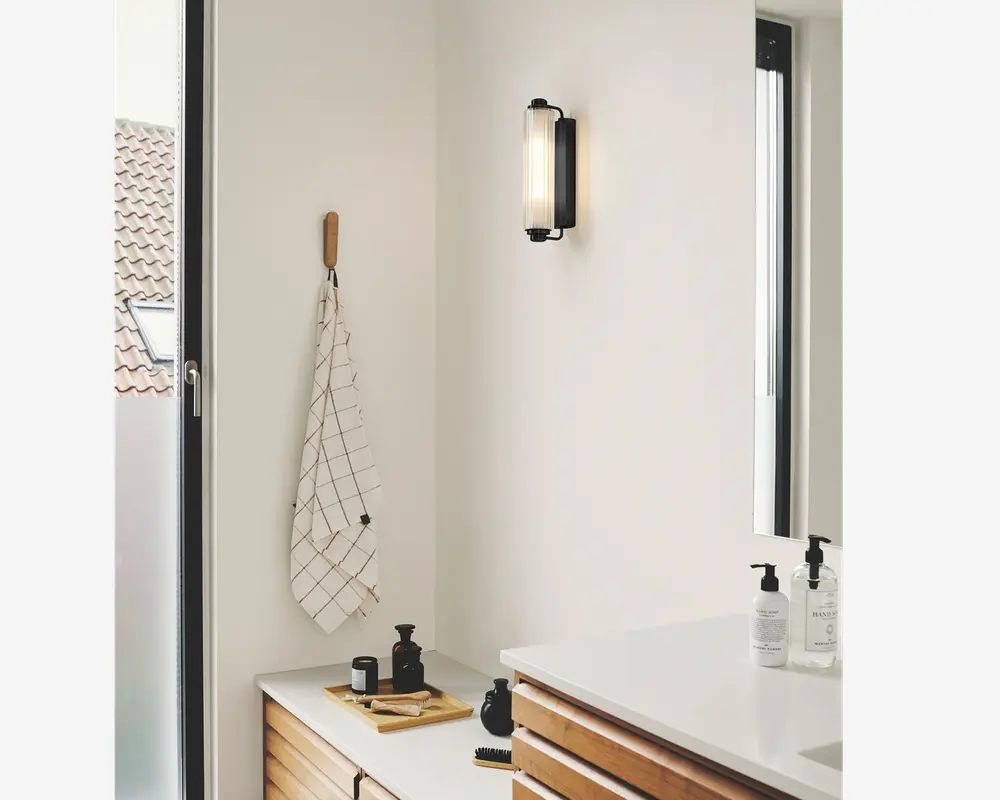 Moderne badeværelse med minimalistisk design, der har en vægmonteret sort og glas væglampe, hvid bordplade med træskuffer og et rektangulært spejl. På bordet står sorte og hvide sæbedispensere, en lille sort krukke og en træbakke med en børste. Et hvidt håndklæde med sort ternet mønster hænger på en træknage. Rummet er lyst med naturligt lys fra et stort vindue.