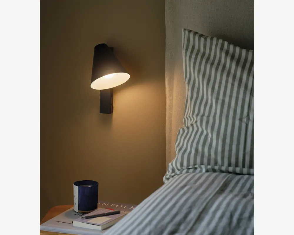 En moderne vægmonteret lampe med en mat sort, konisk lampeskærm, der udsender et varmt, fokuseret lys. Lampen er monteret over et sengebord, hvorpå der står et mørkeblåt krus, en lukket bog og et par briller. Sengen ved siden af lampen har en pude og sengetøj med grønne og hvide lodrette striber, hvilket skaber en hyggelig og indbydende stemning.