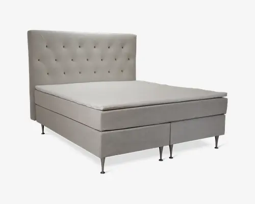 Polstret seng med høj, knappet sengegavl med knapdetaljer. Sengen er betrukket med et lyst beige stof og står på slanke, metalliske ben. Madrassen ser tyk og støttende ud, med en ekstra topmadras for øget komfort. Designet er moderne og elegant, velegnet til nutidige soveværelser.