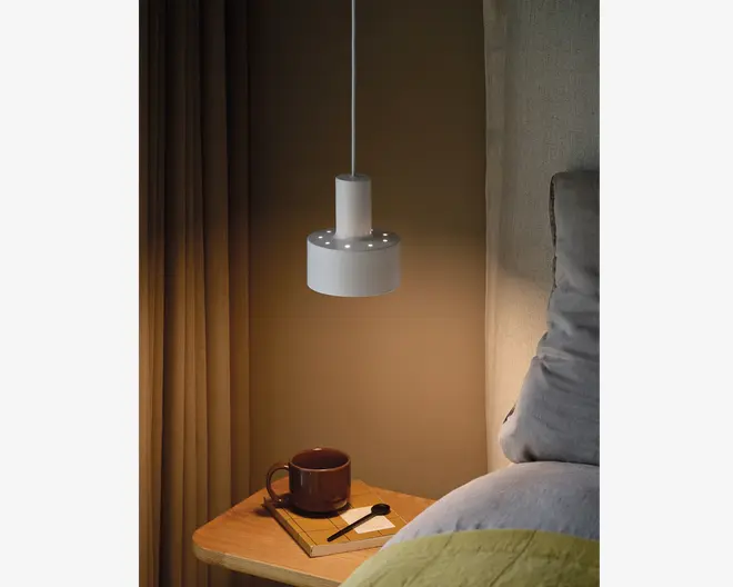 En moderne, cylindrisk hvid pendellampe hænger over et sengebord. Lampen har en glat, mat overflade og en række små, runde huller nær bunden, som lader lyset skinne igennem og skaber en dekorativ effekt. Lampen er ophængt i en tynd, hvid ledning. På sengebordet står et brunt keramikkrus, en lukket notesbog og en kuglepen, hvilket giver et hyggeligt og funktionelt indtryk af soveværelset.