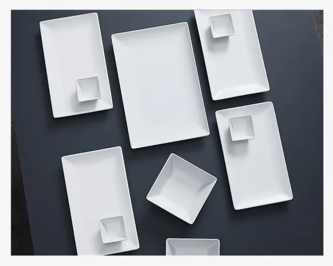 Et udvalg af moderne, minimalistiske hvide serveringsfade i forskellige rektangulære og firkantede former. Sættet indeholder store rektangulære fade og mindre firkantede skåle, alle med rene linjer og blank finish. Fadene er arrangeret på en mørk overflade, hvilket fremhæver deres elegante, moderne design.