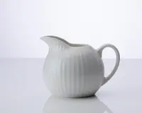 En lille, rund hvid keramikkande med rillet tekstur på overfladen. Den har en bred tud, der gør det nemt at hælde, og et buet håndtag for et behageligt greb. Designet er enkelt og elegant, velegnet til servering af fløde eller mælk.