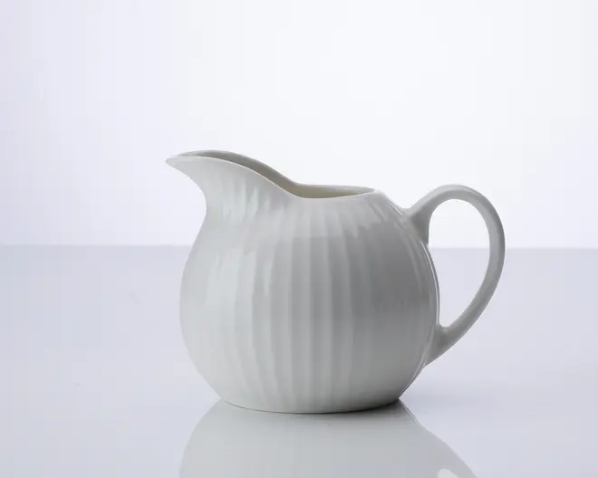 En lille, rund hvid keramikkande med rillet tekstur på overfladen. Den har en bred tud, der gør det nemt at hælde, og et buet håndtag for et behageligt greb. Designet er enkelt og elegant, velegnet til servering af fløde eller mælk.