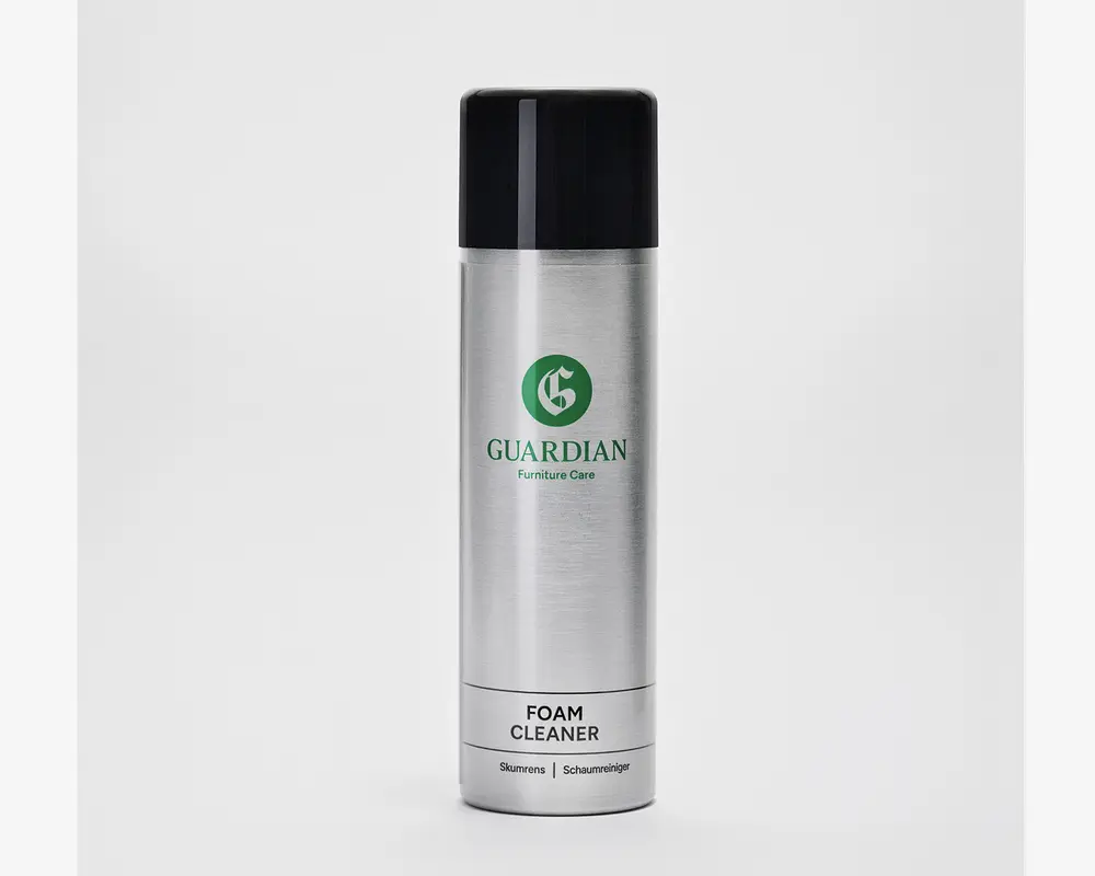 En høj, cylindrisk sølvfarvet spraydåse med sort låg. Dåsen har et grønt logo og mærket 'Guardian Furniture Care' trykt på forsiden. Nederst på dåsen står der 'Foam Cleaner' med fed sort skrift, og der er yderligere tekst i mindre skrift nedenunder. Designet er stilrent og moderne med en metallisk overflade.