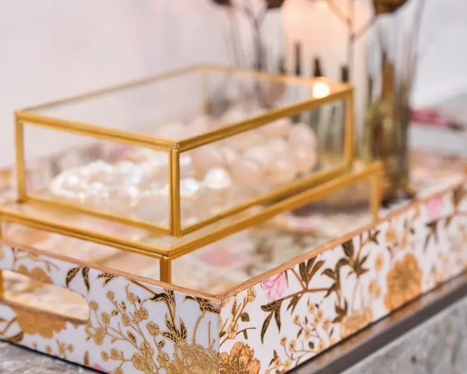 Rektangulær glasæske med guldfarvede metalrammer og hængslet låg, placeret på et dekorativt bakke. Bakken har hvid baggrund med et detaljeret guld- og lyserødt blomstermønster samt udskårne håndtag i siderne. Glasæsken indeholder smykker eller tilbehør, der ligner perler, og det samlede udtryk er elegant og luksuriøst.