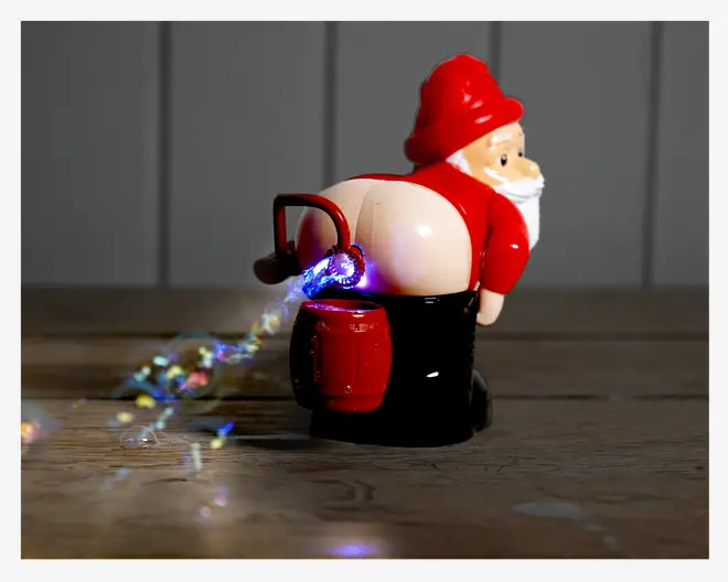 En sjov figur formet som en nisse med rød hue og jakke, der bukker sig forover med bukserne nede og blottet bagdel. Figuren udsender en strøm af farverigt konfetti og lys fra den blottede del, hvilket giver en humoristisk og legende effekt. Nissen holder en rød spand, og det samlede design er lavet for at være morsomt og opsigtsvækkende.
