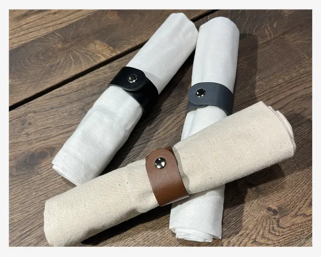 Tre sammenrullede stofservietter holdt sammen af servietringe i læder, hver med en metaltrykknap. Servietringene er i sort, mørkegrå og brun. Servietterne er lavet af et lysefarvet, naturligt stof og ligger på en træoverflade.