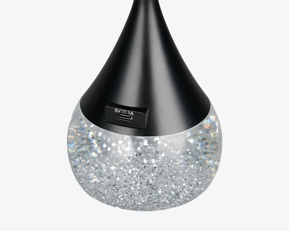 Nærbillede af et produkt med dråbeformet design, hvor den øverste del er sort og mat, og den nederste del er fyldt med glitrende, sølvfarvet materiale. Der er en synlig micro USB-opladningsport mærket '5V⎓1A' på den sorte del, hvilket indikerer, at produktet er genopladeligt. Den glitrende bund antyder en dekorativ eller lysudsendende funktion.