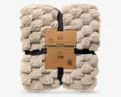 Blød beige plaid med imiteret pels og et kraftigt, fluffy teksturmønster, foldet i en firkant og samlet med et brunt papband.