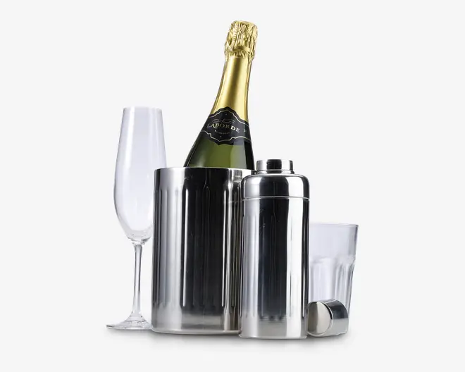 Et sæt barudstyr bestående af et højt, klart champagneglas, en isspand i rustfrit stål med en flaske champagne med gyldent folie, en matchende cocktailshaker i rustfrit stål med aftageligt låg og et klart glas tumbler. Produkterne har en glat, poleret overflade, hvilket giver et moderne og elegant udtryk.