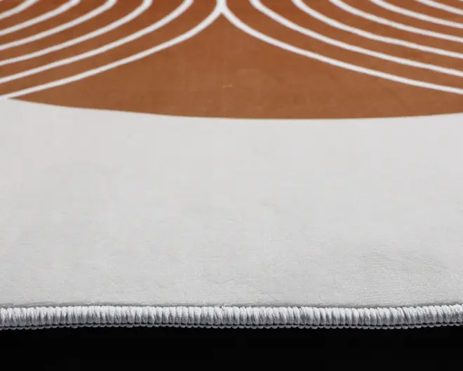 Nærbillede af et tæppe, der viser en tyk, blød tekstur med pænt syede kanter. Tæppet har et moderne design med et buet hvidt område og et brunt felt dekoreret med hvide, jævnt fordelte, buede linjer. Syningen langs kanten fremstår holdbar og veludført.