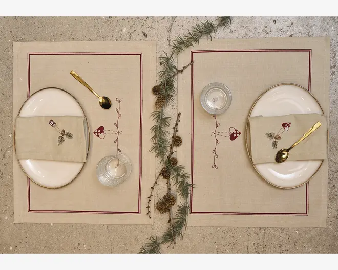 To beige borddækninger er vist side om side på en stengulvsoverflade, hver med en dækkeserviet med rød kant, oval hvid tallerken, foldet matchende serviet og en gylden ske. Små klare glas står ved tallerkenerne, og en fyrregren med kogler er lagt mellem opdækningerne for et rustikt udtryk.