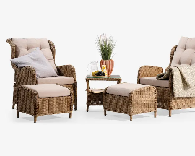 Sættet består af to høje lænestole med tykke, polstrede beige hynder, to matchende skamler og et lille sidebord. Møblerne er lavet af flettet rattan i en naturlig brun farve. Den ene stol har en hvid og en grå pude, mens den anden har et lysegrønt tæppe lagt over armlænet. Sidebordet har en dekorativ vase med kunstigt græs, et glas vand med citron og en lille tallerken med druer. Udtrykket er hyggeligt og indbydende, velegnet til både indendørs og udendørs brug.