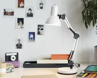 Justerbar skrivebordslampe med hvid metalskærm og fod, samt sort og sølvfarvet ledarm for fleksibel placering. Lampen har et moderne, minimalistisk design og står på et arbejdsbord med farveblyanter, notesbøger og dekorative genstande i nærheden.