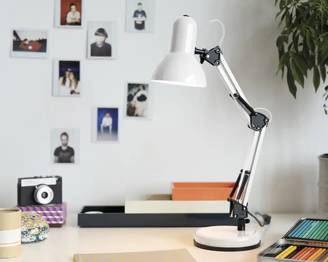 Justerbar skrivebordslampe med hvid metalskærm og fod, samt sort og sølvfarvet ledarm for fleksibel placering. Lampen har et moderne, minimalistisk design og står på et arbejdsbord med farveblyanter, notesbøger og dekorative genstande i nærheden.