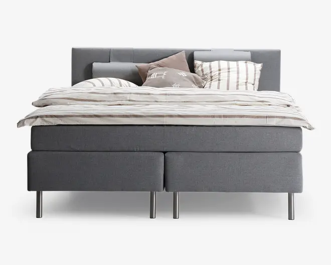 En moderne seng med gråt polstret stel og sengegavl, der har rene linjer og et minimalistisk design. Sengen er redt med et lysebeige og hvidt stribet dynebetræk og matchende puder samt ekstra pyntepuder i neutrale nuancer. Sengen står på slanke, metalliske ben, hvilket giver et moderne udtryk.