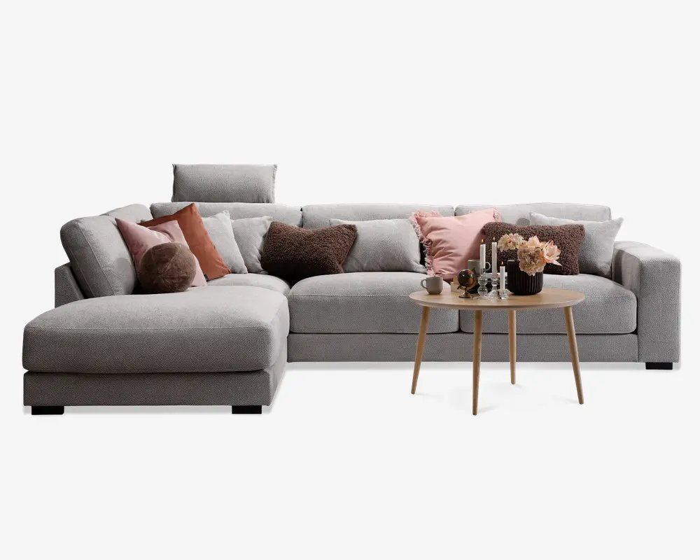 Moderne hjørnesofa i lysegråt stof med chaiselong til venstre. Sofaen har flere bløde puder i forskellige farver, herunder brun, pink og hvid, hvilket giver et hyggeligt og indbydende udtryk. Foran sofaen står et rundt sofabord i træ, dekoreret med stearinlys, en lille vase med blomster og andre dekorative genstande. Sofaen har rene linjer og et moderne design, der passer til en stilfuld stue.