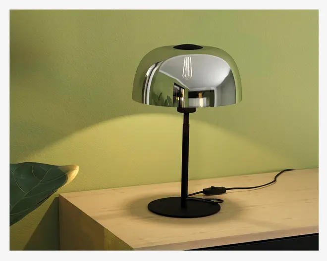 En moderne bordlampe med en blank, reflekterende metalskærm i kuppelform og en slank sort stander. Lampen har en rund sort fod og en synlig sort ledning med en afbryder på ledningen. Den metalliske skærm spejler omgivelserne, og lampen udsender et varmt, blødt lys, der giver en mild glød på væggen og overfladen.