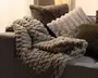 Et tykt, blødt plaid med en kraftig, løkket struktur, lagt over en sofa og kombineret med flere bløde pyntepuder i neutrale farver.