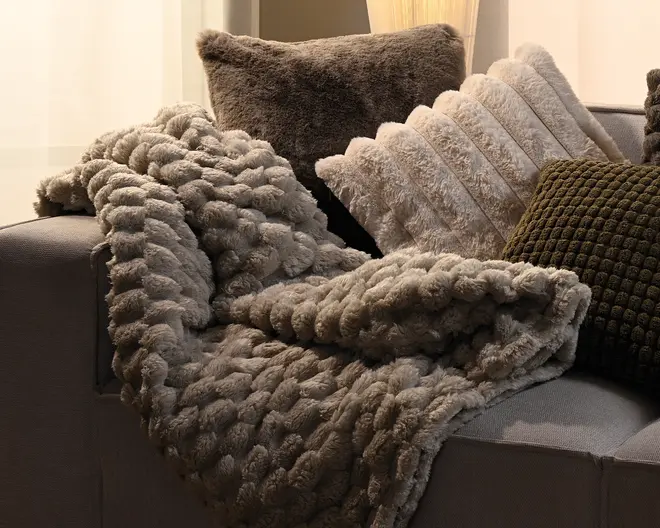Et tykt, blødt plaid med en kraftig, løkket struktur, lagt over en sofa og kombineret med flere bløde pyntepuder i neutrale farver.