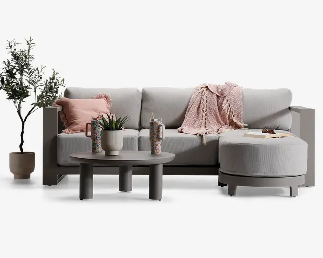 Moderne hjørnesofa med lysegråt betræk, brede armlæn og bløde ryghynder. Sofaen har en rund chaiselong til højre. Den er pyntet med et tekstureret lyserødt tæppe, en matchende lyserød pude og en dekorativ plante på venstre armlæn. Foran sofaen står et rundt sofabord med mat finish, hvorpå der står en potteplante og to mønstrede krus. Udtrykket er hyggeligt og moderne med bløde, neutrale farver og diskrete dekorative detaljer.