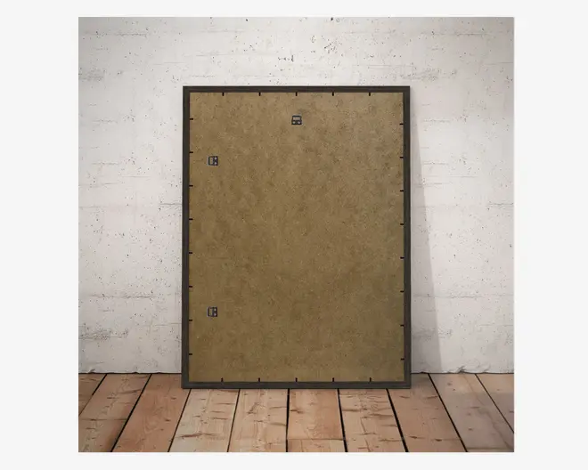Rektangulært objekt med sort ramme og brun, tekstureret bagplade, der har tre metalophængsbeslag til vægmontering. Bagpladen er fastgjort med flere sorte klips langs kanterne. Objektet står oprejst mod en hvid, tekstureret væg og et trægulv.