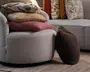 Lysegråt, afrundet møbel med blødt tekstureret betræk, stablede pyntepuder i beige, rosa og mønstrede betræk samt en mørk, fluffy pyntepude, der læner sig mod siden.