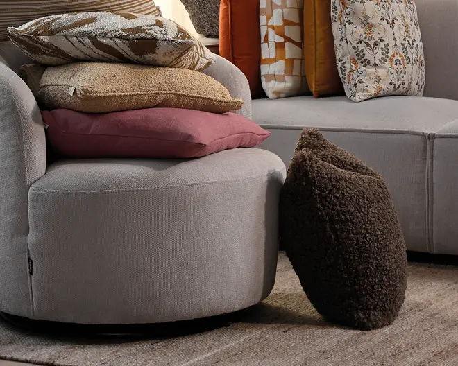 Lysegråt, afrundet møbel med blødt tekstureret betræk, stablede pyntepuder i beige, rosa og mønstrede betræk samt en mørk, fluffy pyntepude, der læner sig mod siden.