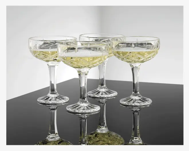 Sæt med fire klare coupeglas med et vintage-inspireret mønster i slebet glas på skål og fod. Hvert glas er fyldt med en lys mousserende drik, som fremhæver det indviklede geometriske design. Glassene har korte, solide stilke og brede, lave skåle, ideelle til servering af champagne eller cocktails. Den reflekterende overflade under glassene forstærker det elegante udtryk.