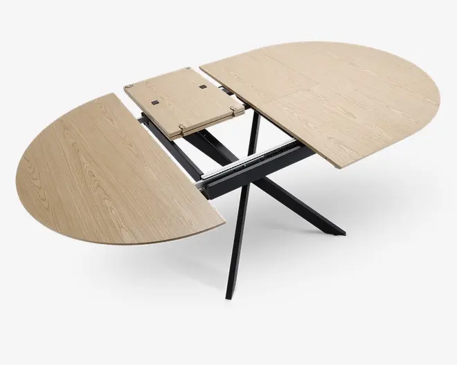 Ovalformet bord med lys træfinish, vist i udtrukket position med en midterplade indsat for at øge længden. Bordpladen har en glat overflade med synlige træårer. Bordet har en sort metal krydsbenet base, som giver stabilitet og et moderne udtryk. Udtræksmekanismen er synlig og viser, hvordan bordet kan justeres for ekstra siddepladser eller plads.