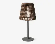 Bordlampe med en unik lampeskærm i bølgeformet design, lavet af bronzefarvede metalstrimler. Skærmen har et åbent, luftigt udtryk, der lader lyset passere gennem mellemrum. Lampen har en enkel, rund fod og en lige, matgrå stang, hvilket giver et moderne og kunstnerisk udseende.