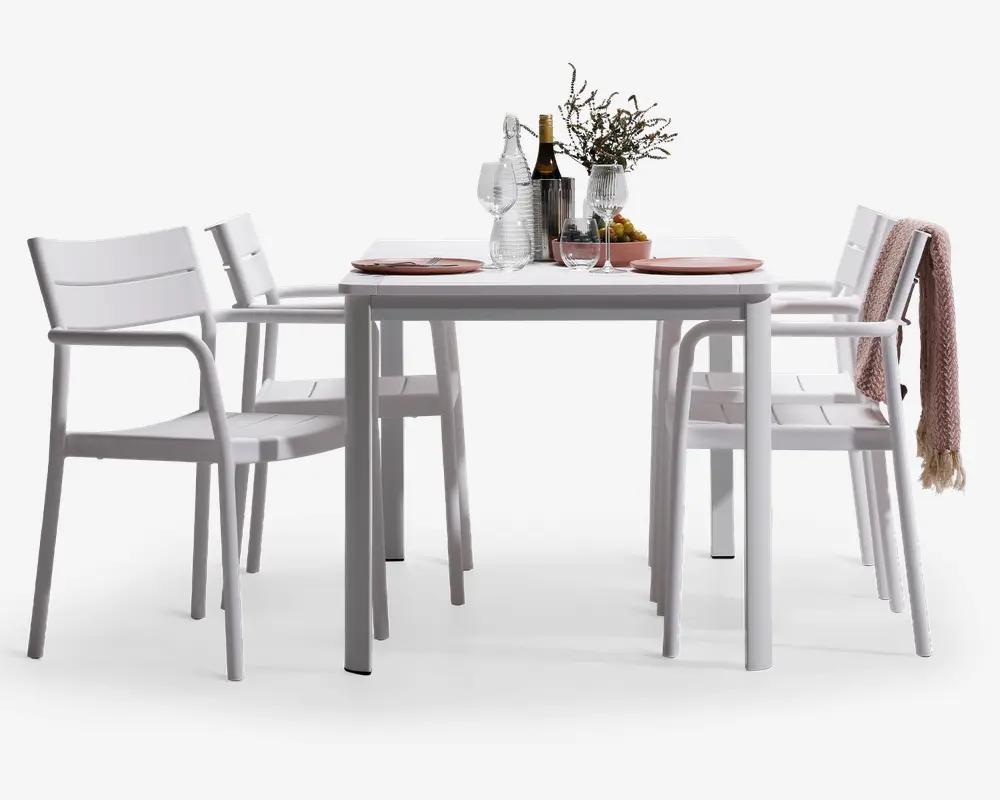 Et moderne udendørs spisesæt bestående af et rektangulært hvidt bord med fire matchende hvide stole. Stolene har armlæn samt lameller i ryg og sæde for komfort og ventilation. Bordet er dækket med tallerkener, glas, en karaffel, vinflasker og en dekorativ vase med blomster. Et tekstureret tæppe er lagt over ryggen på en af stolene, hvilket giver et hyggeligt præg. Designet er stilrent og minimalistisk, velegnet til moderne udendørsområder.
