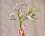 En hånd holder en lille buket kunstige blomster med fine lyse lilla og hvide blomster. Stilkene er grønne med en blanding af lange, slanke blade og mindre løv. Blomsterne har et realistisk udseende, hvor nogle kronblade er let krøllede, og andre er mere kompakte. Arrangementet fremstår luftigt og naturligt, velegnet til dekoration.