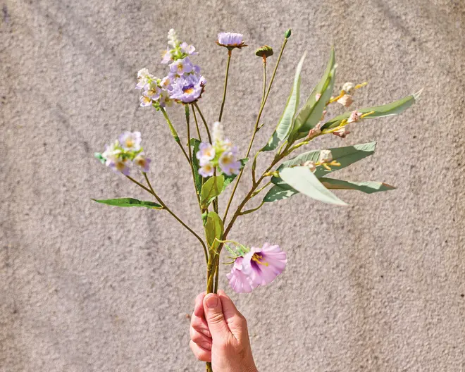 En hånd holder en lille buket kunstige blomster med fine lyse lilla og hvide blomster. Stilkene er grønne med en blanding af lange, slanke blade og mindre løv. Blomsterne har et realistisk udseende, hvor nogle kronblade er let krøllede, og andre er mere kompakte. Arrangementet fremstår luftigt og naturligt, velegnet til dekoration.