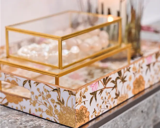 Rektangulært dekorativt bakke med udskårne håndtag, hvid baggrund og guld- samt lyserøde blomsterdekorationer. Ovenpå bakken står en glaskasse med guldfarvede metalrammer, som indeholder smykker af perler og ædelsten. Udtrykket er elegant og luksuriøst, velegnet til organisering eller udstilling af accessories.