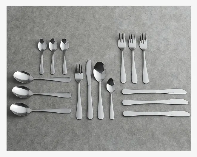 Et sæt bestik i rustfrit stål arrangeret på en grå overflade, bestående af tre små teskeer, tre store spiseskeer, tre middagsgafler, tre middagsknive, én suppeske og én dessertske. Hvert stykke har et enkelt, moderne design med en glat, poleret finish og let afrundede håndtag.