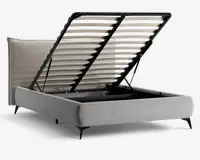 Polstret sengestel i lysebeige stof med en høj, polstret sengegavl, der har et diskret geometrisk mønster. Sengen har en opklappelig lamelbund med sort metalramme, som giver adgang til et rummeligt opbevaringsrum under sengen. Sengen står på korte, skrå sorte ben, hvilket giver et moderne udtryk.