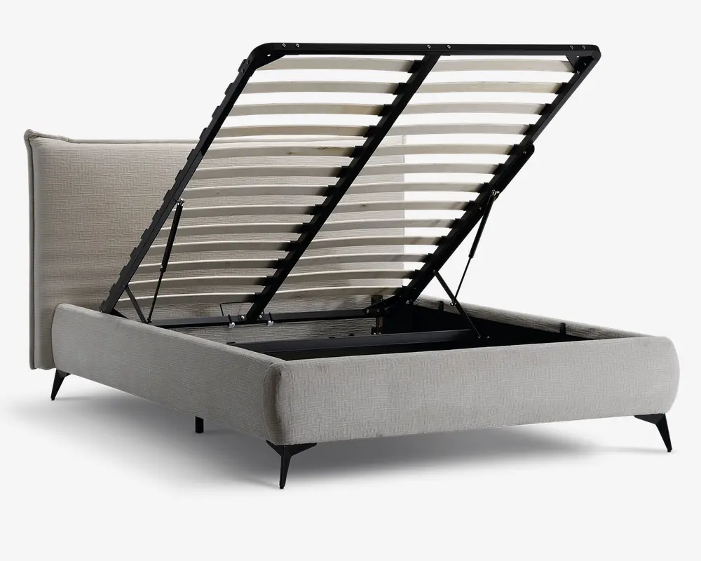 Polstret sengestel i lysebeige stof med en høj, polstret sengegavl, der har et diskret geometrisk mønster. Sengen har en opklappelig lamelbund med sort metalramme, som giver adgang til et rummeligt opbevaringsrum under sengen. Sengen står på korte, skrå sorte ben, hvilket giver et moderne udtryk.