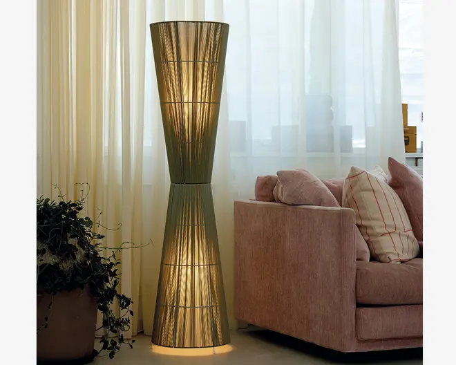 Høj gulvlampe med timeglasform og en vævet, halvgennemsigtig lampeskærm, der spreder et varmt lys. Lampen har et symmetrisk design med en smal midte og bredere top og bund. Skærmen består af tynde, lodrette tråde, hvilket giver et tekstureret udseende. Lampen står på en rund fod og udsender et blødt, stemningsfuldt lys, velegnet til moderne eller nutidige indretninger.