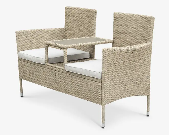 Udendørs bænk med to siddepladser, fremstillet af lysebeige flettet rattan med et hævet bord i midten mellem sæderne. Bænken har høje ryglæn og armlæn på begge sider, og hvert sæde har en aftagelig, tyk hvid hynde for ekstra komfort. Bordet er integreret i rammen og giver en praktisk overflade til drikkevarer eller småting.