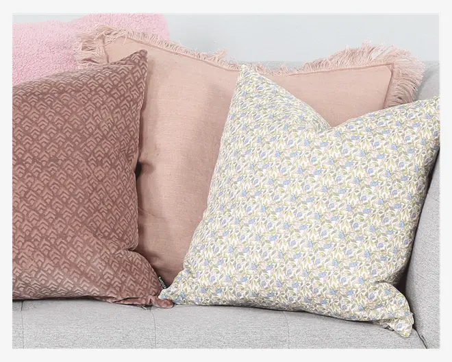 Tre dekorative puder er arrangeret på en lysegrå sofa. Den første pude til venstre har et tekstureret geometrisk mønster i en afdæmpet mauve farve. Den midterste pude er ensfarvet lyserød med blødt stof og frynset kant. Puden til højre har et fint blomstermønster med små blå og gule blomster på en lys baggrund, hvilket giver et blødt og indbydende udtryk.