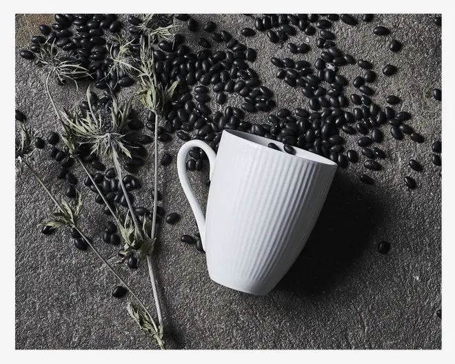 En hvid keramikkop med lodret rillet tekstur og et buet håndtag. Koppen ligger på en ru, stenlignende overflade omgivet af spredte sorte bønner og tørrede, fine plantestængler. Designet er minimalistisk og moderne med en let tilspidset form.