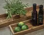 Rektangulære træbakker med naturlig finish, hvor den ene indeholder en grøn krydderurt i potte indpakket i brunt papir, og den anden har tre friske limefrugter samt to flasker – én med rødvinseddike og én med balsamicoeddike. Bakkerne har forhøjede kanter og et enkelt, rustikt design.
