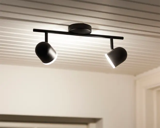 Loftmonteret lampearmatur med et moderne, minimalistisk design. Den har en sort metalstang med to justerbare cylindriske spotlys, som giver mulighed for retningsbestemt belysning. Armaturet er fastgjort til loftet med en rund base, og spotlysene har en mat sort finish med hvide indersider for bedre lysrefleksion.